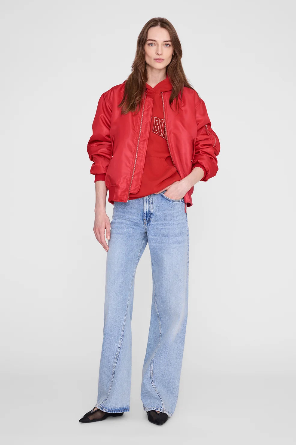 A model stands against a light gray studio background wearing a red satin bomber jacket over a red top, light-wash wide-leg jeans, and black pointed flats.
