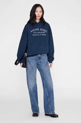 Person wearing a navy hoodie with 'Anine Bing' logo and blue jeans on a white background