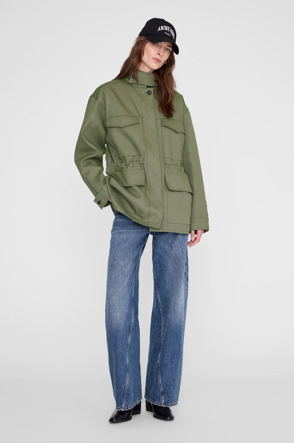 A model wearing an olive green utility jacket, blue wide-leg jeans, and a black baseball cap with white text, standing against a light gray studio background.