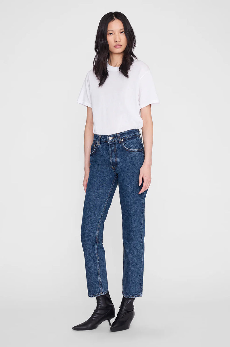 A model wearing a white short-sleeve t-shirt, blue high-waisted straight-leg jeans, and black pointed-toe ankle boots, standing against a light gray studio background.