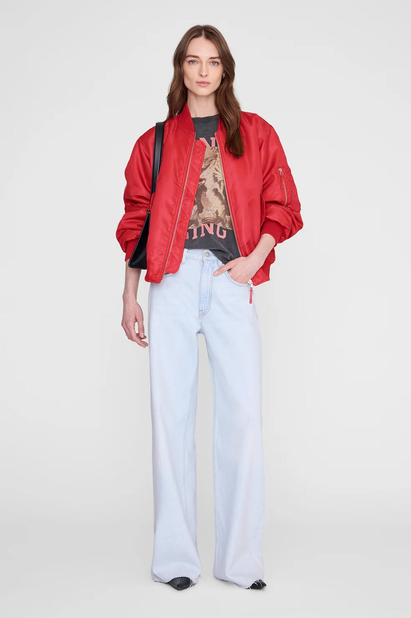 A model stands against a light gray studio background wearing a bright red bomber jacket over a dark graphic T-shirt, paired with very light blue wide-leg jeans and black pointed shoes.