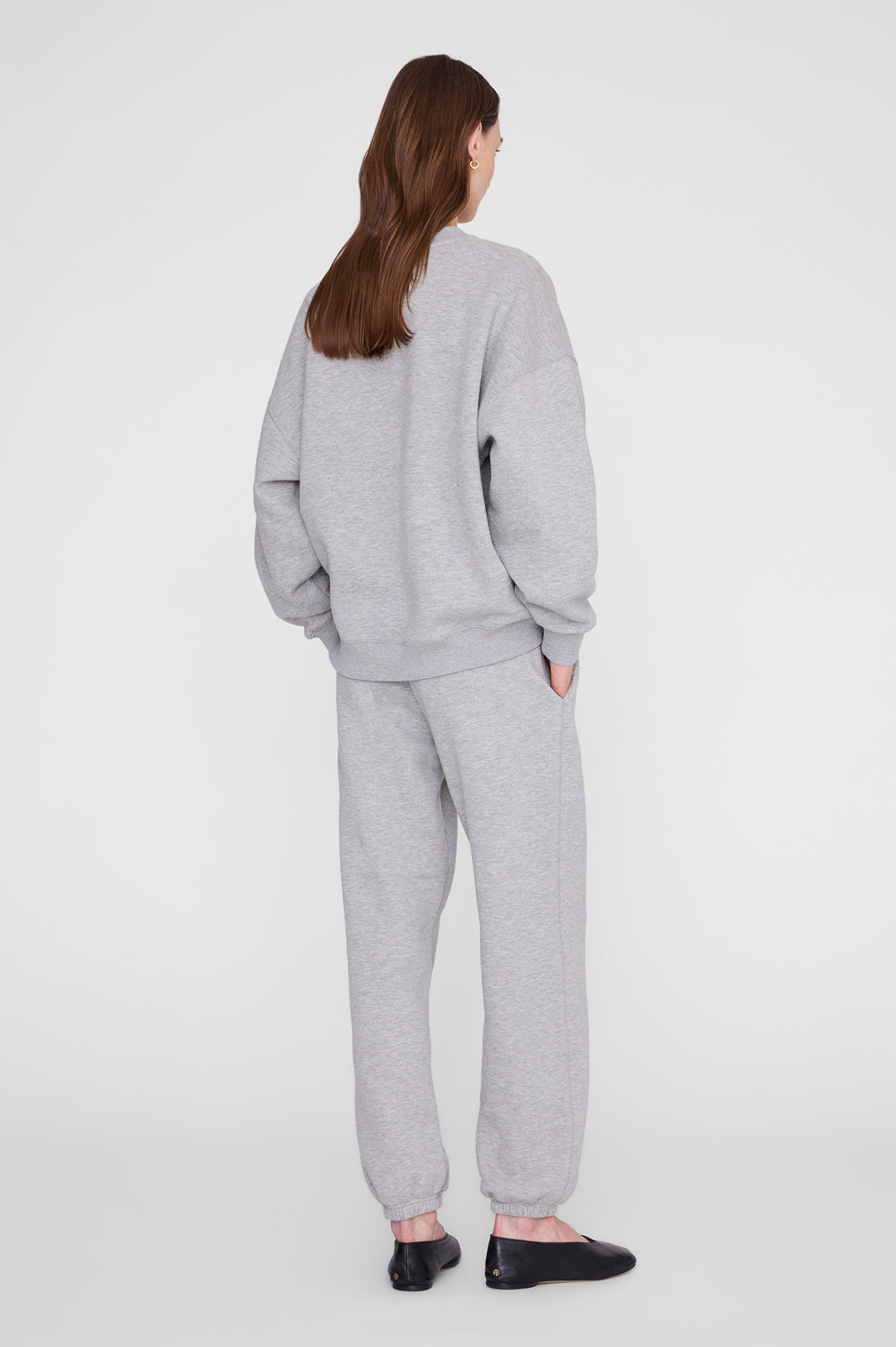 A woman viewed from the back wearing a light gray heather sweatshirt and matching jogger pants, paired with black flats, against a plain light gray studio background.