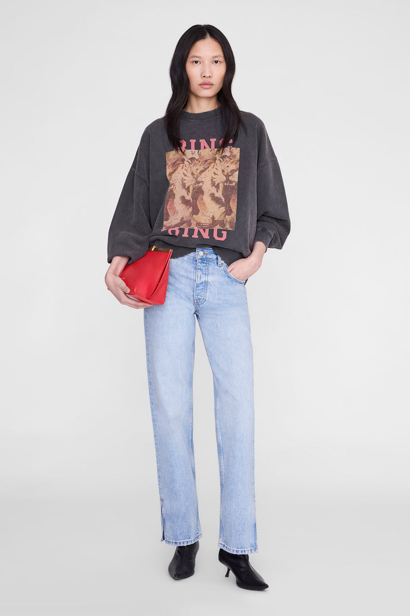 A model stands against a light gray studio background wearing a dark charcoal graphic sweatshirt, light-wash blue jeans, black pointed boots, and holding a bright red clutch.