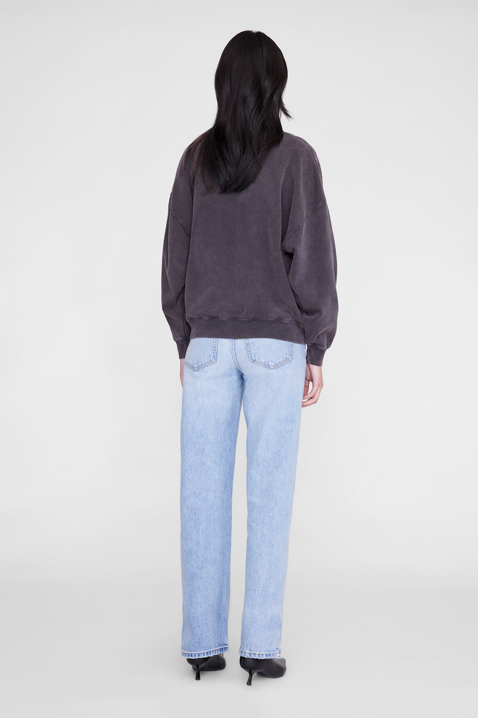 A model shown from the back wearing a dark charcoal gray oversized crewneck sweatshirt and light-wash blue straight-leg jeans, paired with black heels, against a plain light gray studio background.
