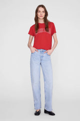 A woman modeling a red graphic t-shirt and light-wash blue jeans against a plain light gray studio background.