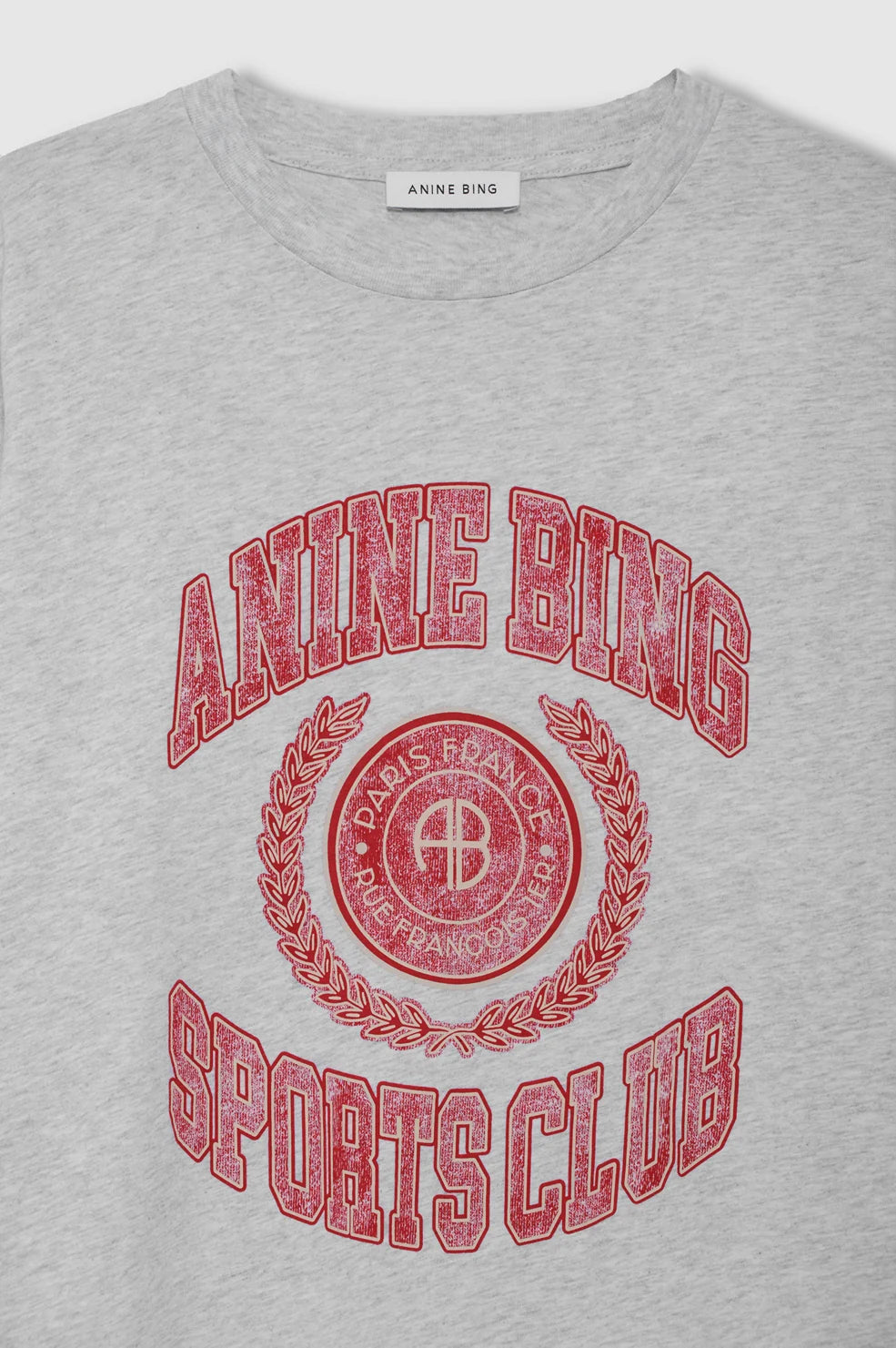 A close-up of a light heather gray t-shirt featuring a distressed red graphic print that reads 'ANINE BING SPORTS CLUB' with a circular crest and laurel wreath. The neck label clearly shows the brand 'ANINE BING'.