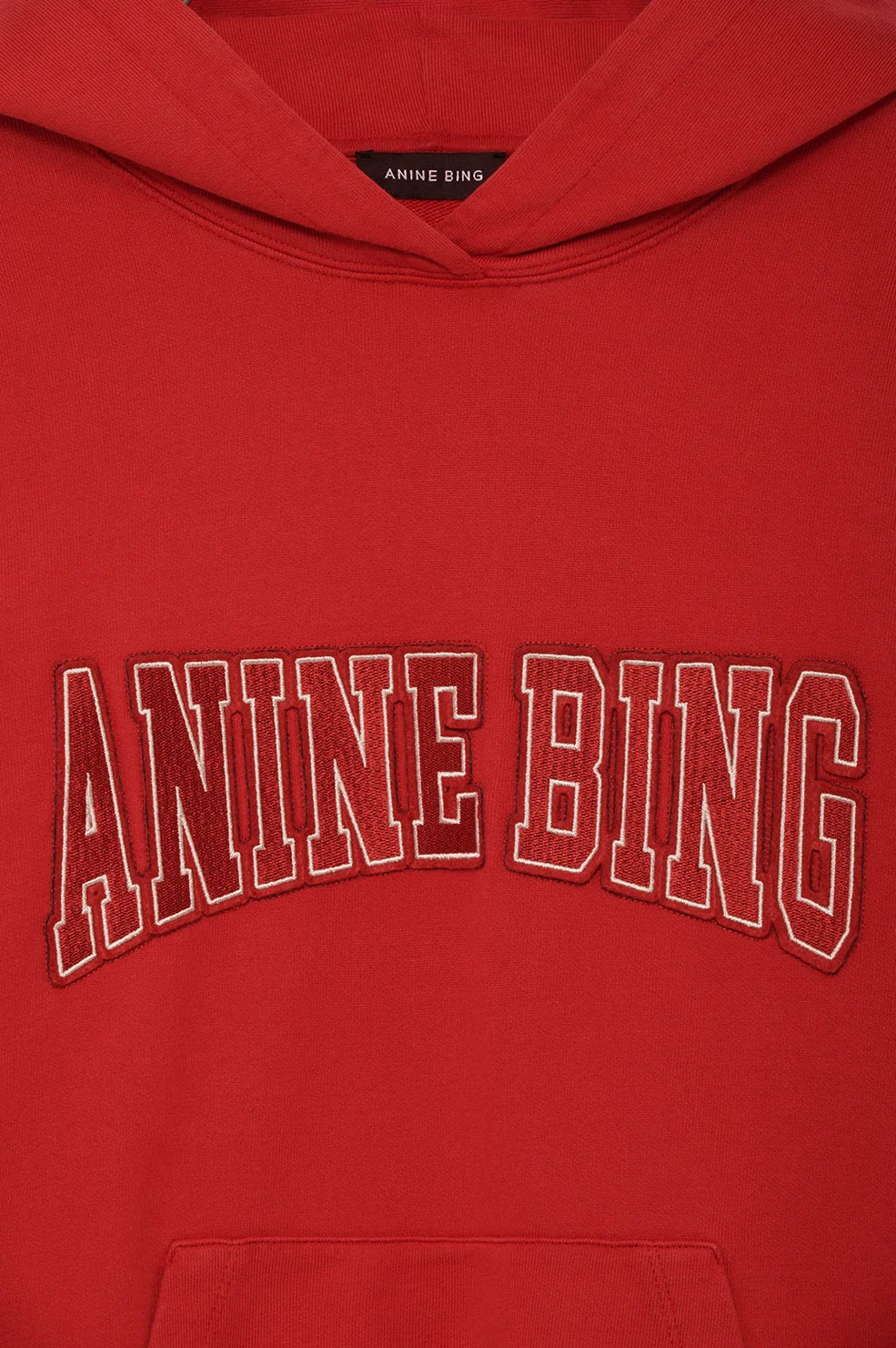 A close-up of a red hoodie featuring the brand name 'ANINE BING' embroidered in arched varsity block letters across the chest, with a matching red inner neck label also visible.