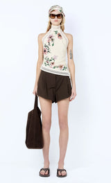 Woman wearing a floral sleeveless top and brown shorts, holding a bag on a white background