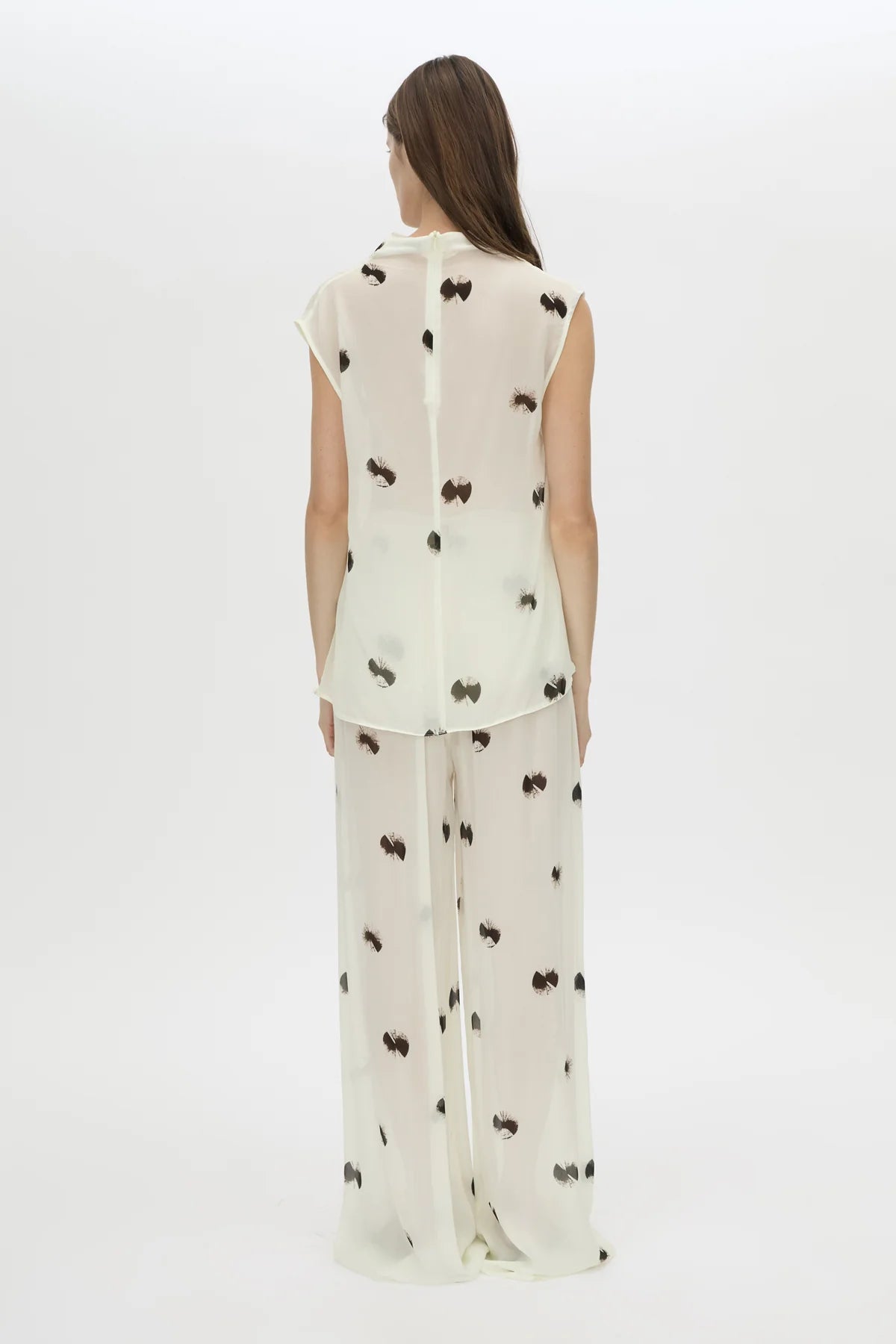 A woman viewed from the back wearing a sheer white sleeveless top and matching wide-leg pants with a scattered black bow print, set against a plain white studio background.
