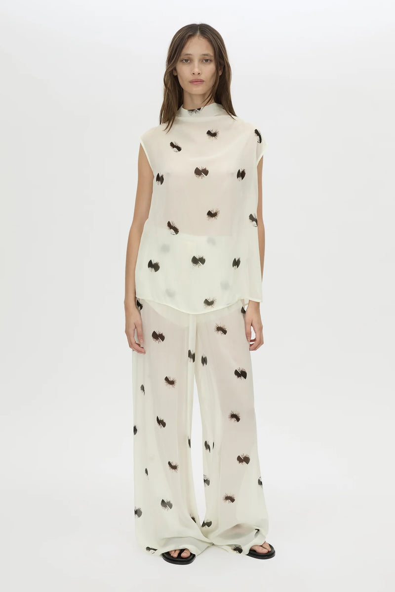 A model wearing a matching two-piece sheer white set with black embroidered bow motifs, consisting of a mock-neck sleeveless top and wide-leg trousers, photographed against a plain white studio background.