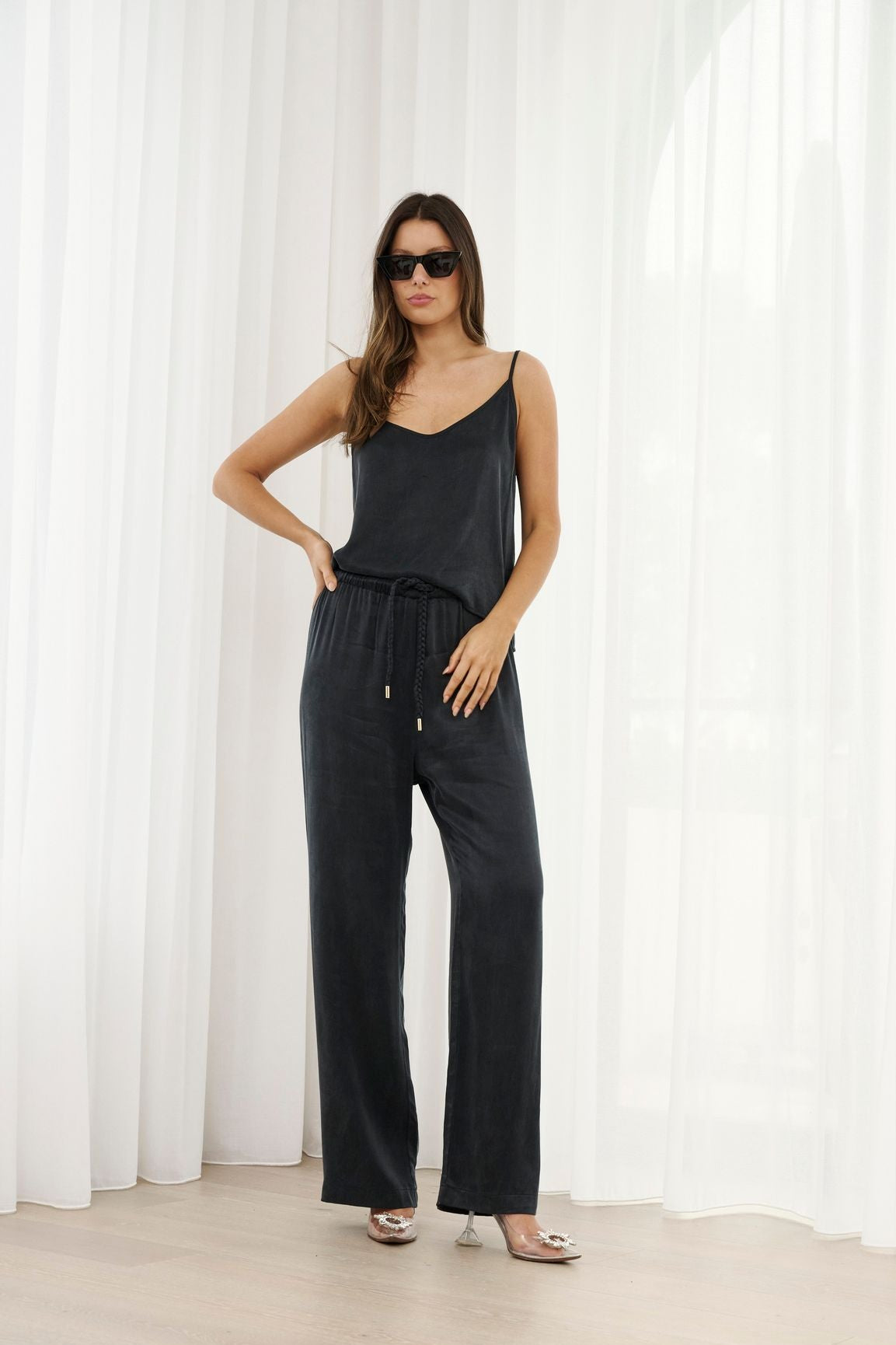 Woman wearing a black sleeveless top and wide-leg pants against a white curtain background