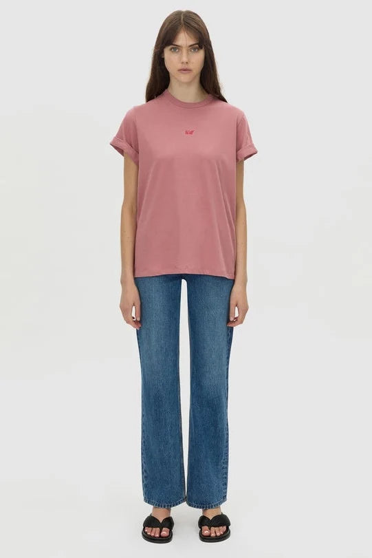 A model stands against a light gray studio background wearing a dusty rose short-sleeve t-shirt with a small red embroidered logo on the chest, blue jeans, and black slide sandals.