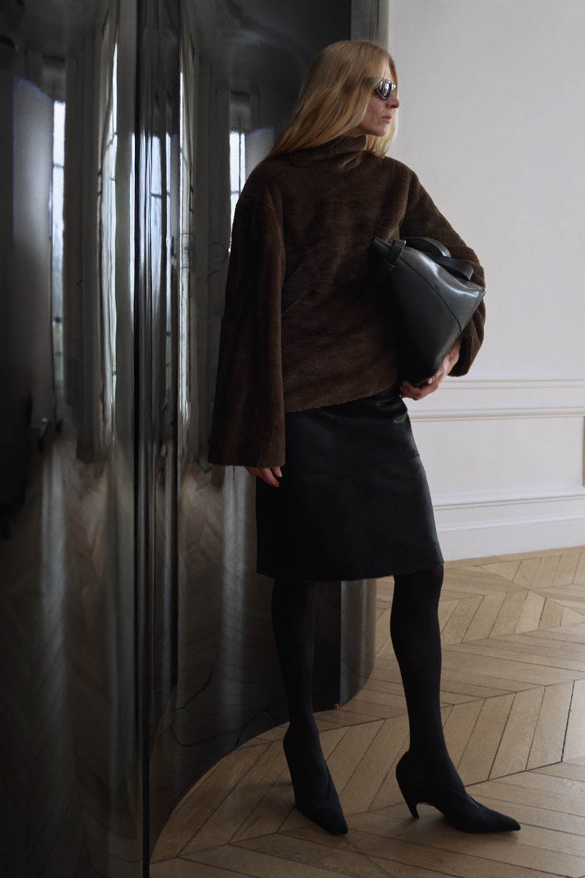 Woman in a brown coat and black skirt standing next to a refrigerator.