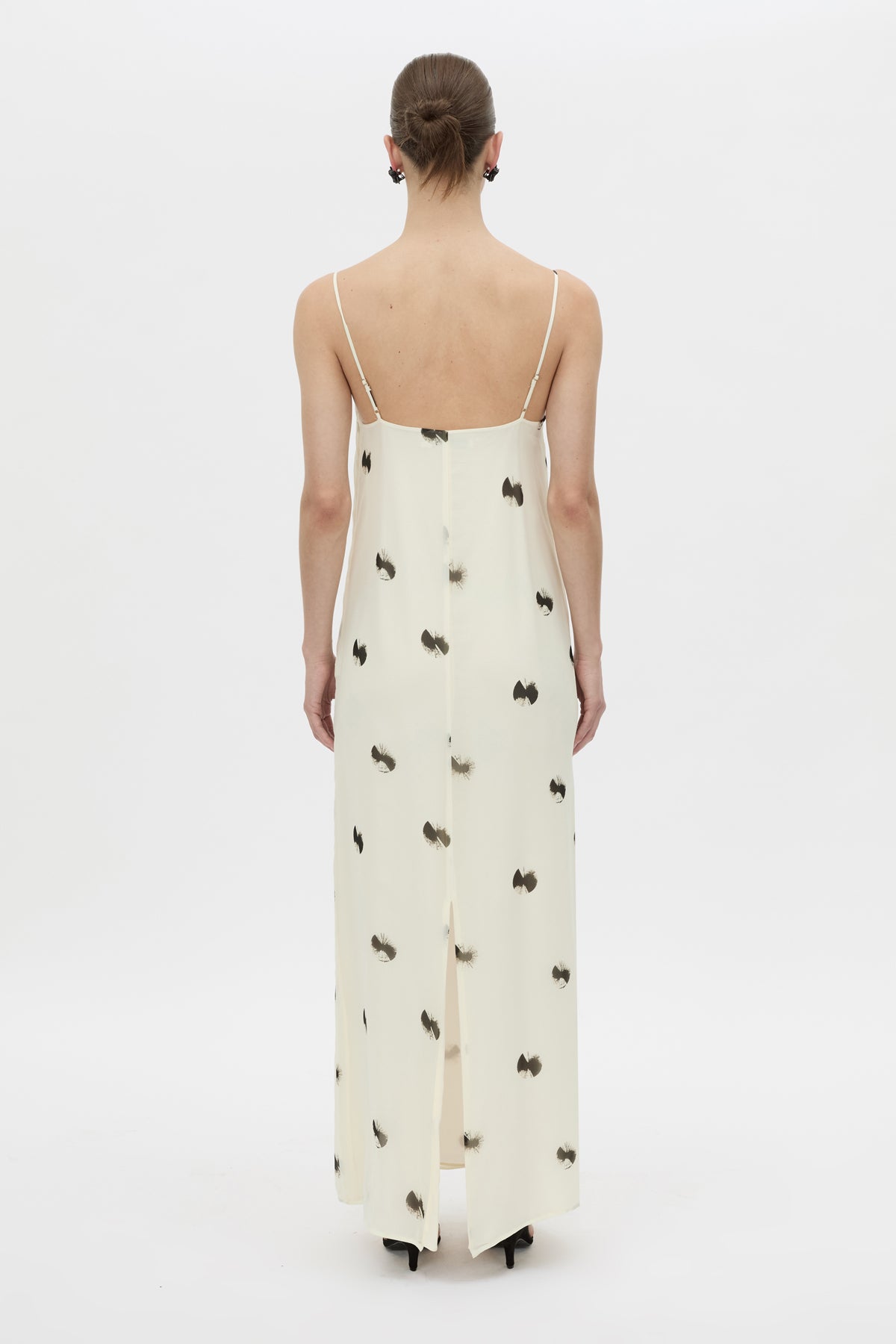 A woman viewed from the back wearing a cream maxi dress with thin straps and a scattered black bow tie print, set against a plain white studio background.