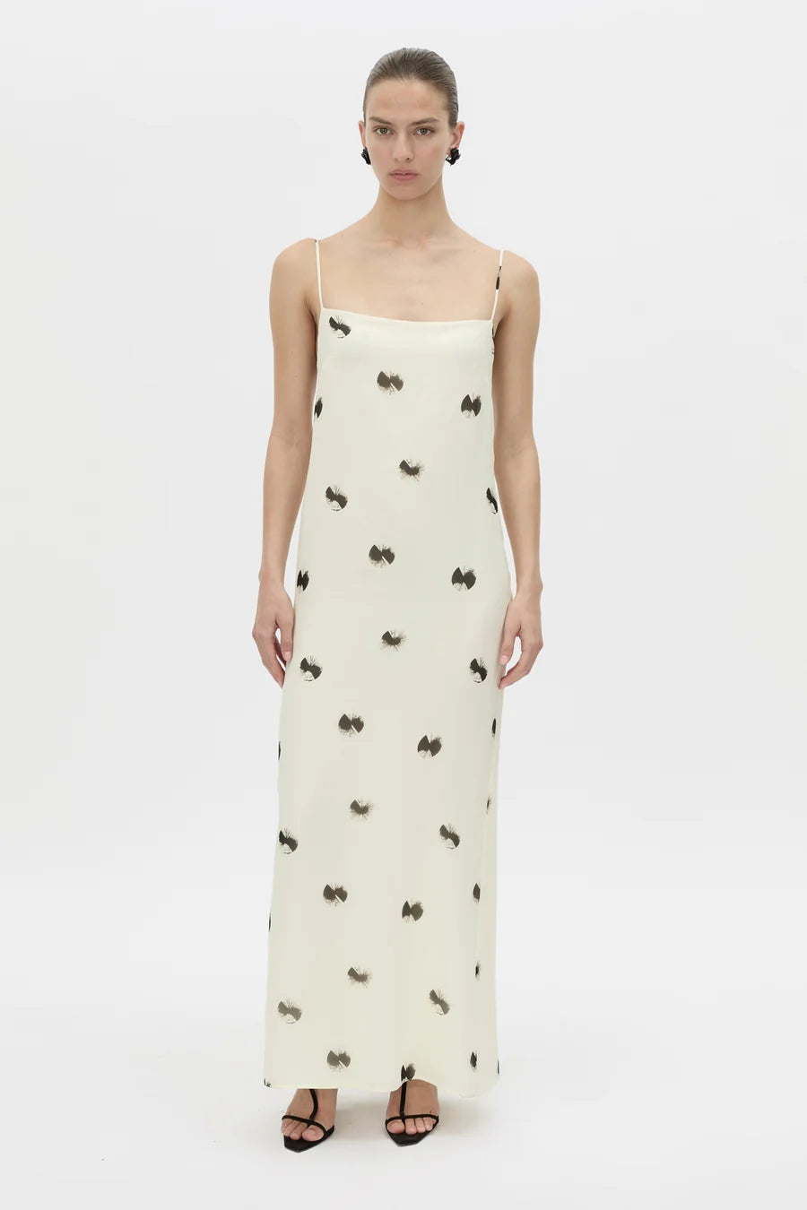 A model wearing a cream-colored maxi slip dress with thin straps and a black bow pattern, paired with black strappy sandals, against a plain white studio background.