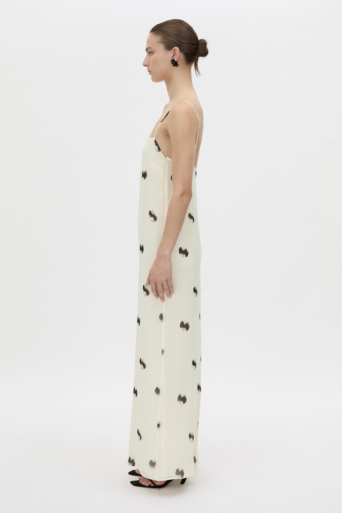 A woman stands in profile wearing a long cream maxi dress with a scattered black butterfly print, photographed against a seamless white studio background.