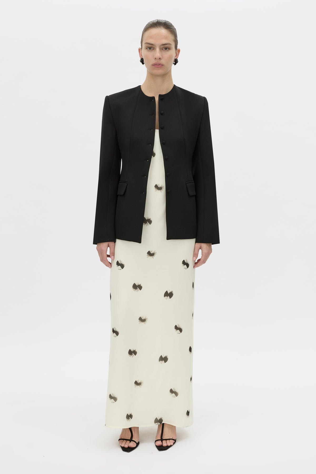 A model stands against a plain white studio background wearing a black cropped jacket over a long cream dress with small dark bow motifs, paired with black strappy heels.