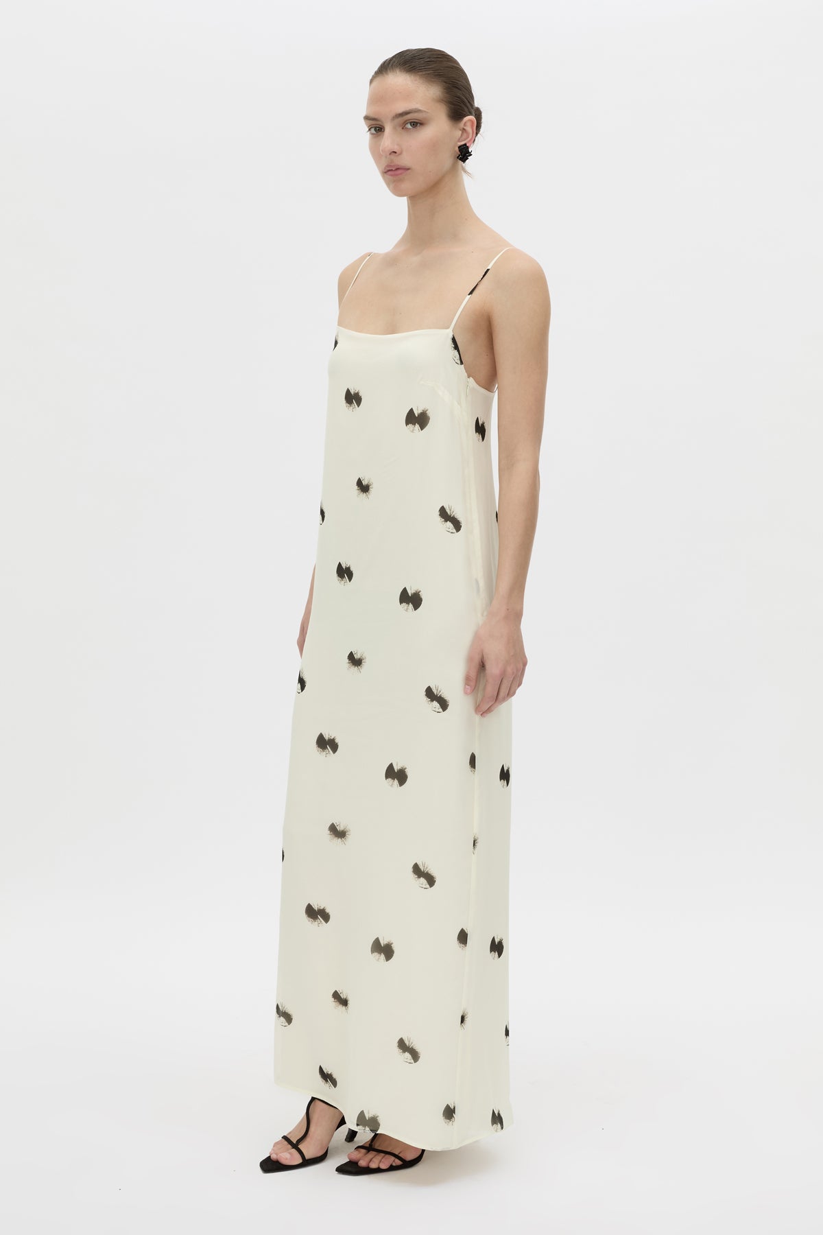 A model wearing a long cream-colored spaghetti-strap dress with a black bow pattern, styled with black strappy sandals against a plain white studio background.