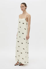 A model wearing a long cream-colored spaghetti-strap dress with a black bow pattern, styled with black strappy sandals against a plain white studio background.