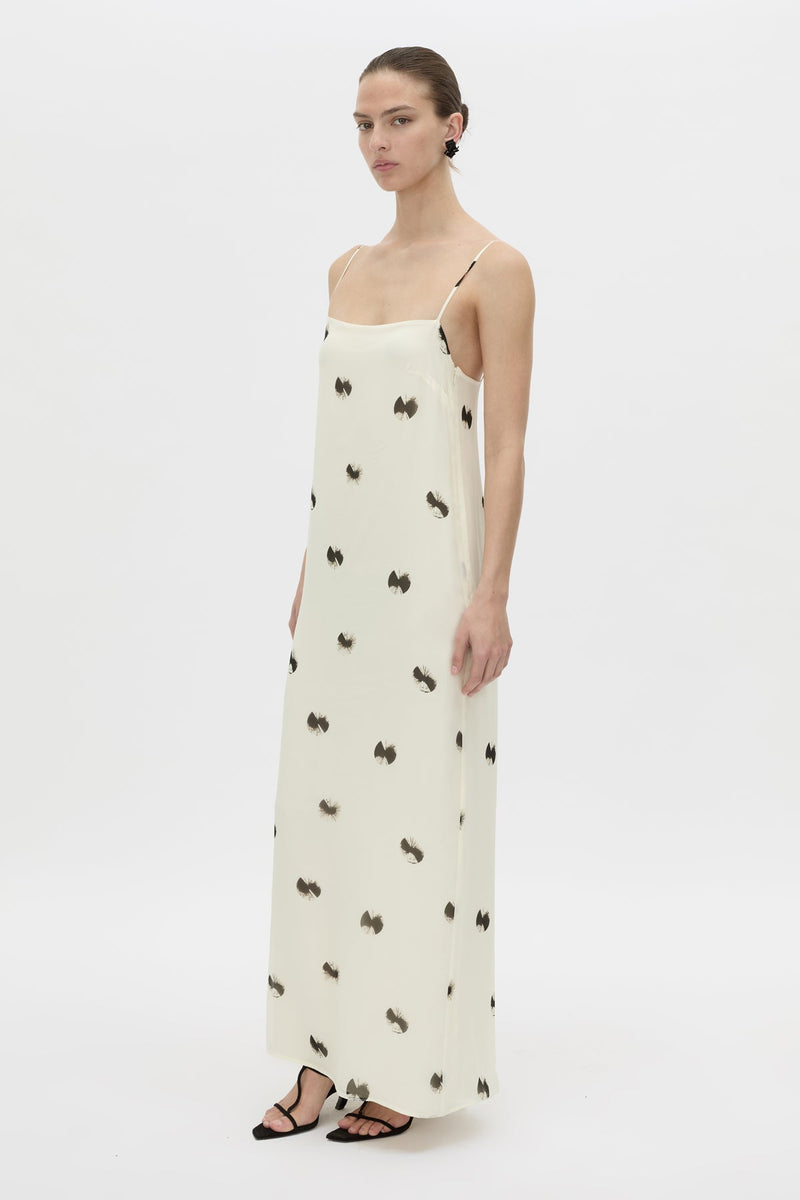A model wearing a long cream-colored spaghetti-strap dress with a black bow pattern, styled with black strappy sandals against a plain white studio background.