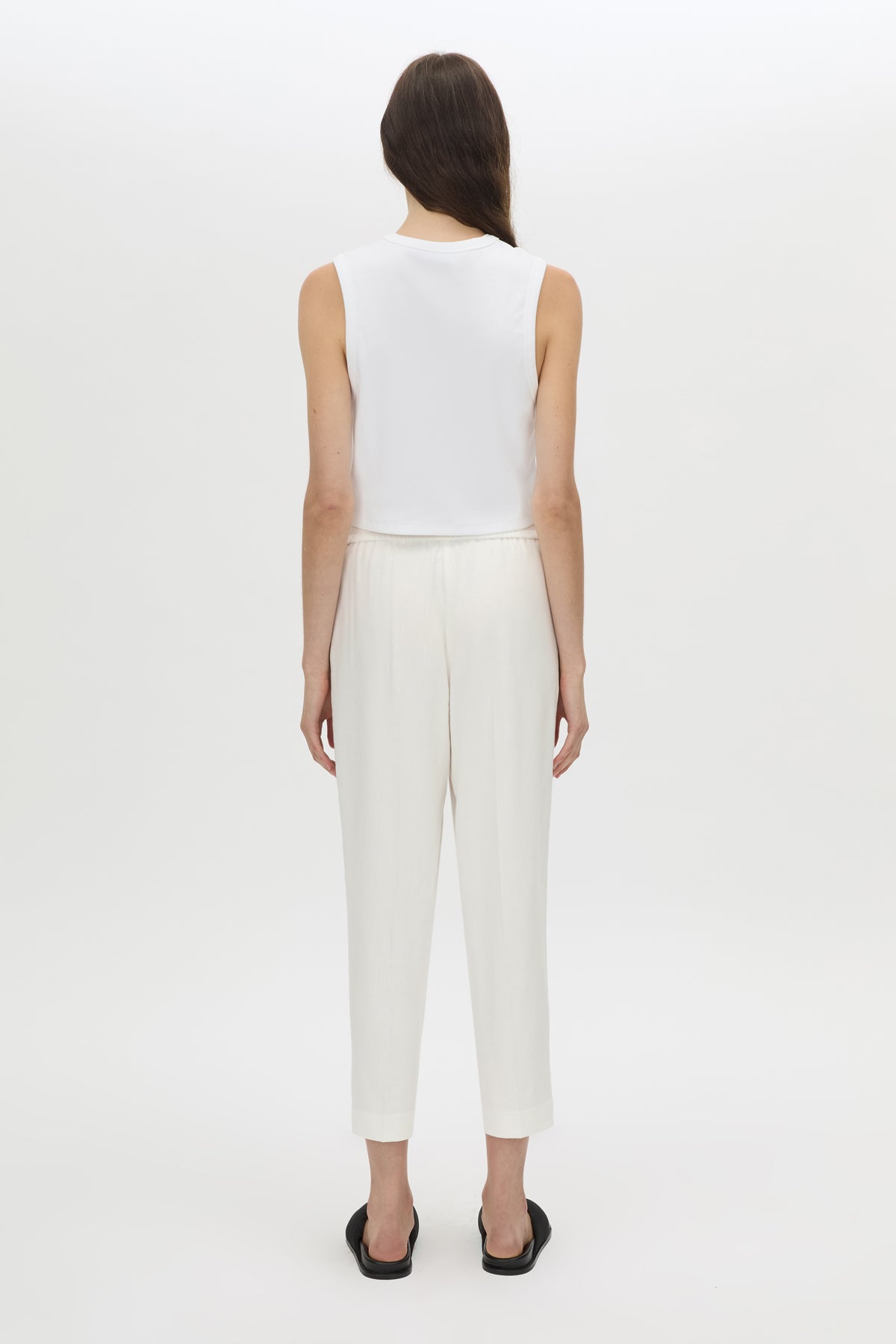 A woman viewed from the back wearing a white sleeveless top and white cropped trousers, standing against a plain light gray studio background.