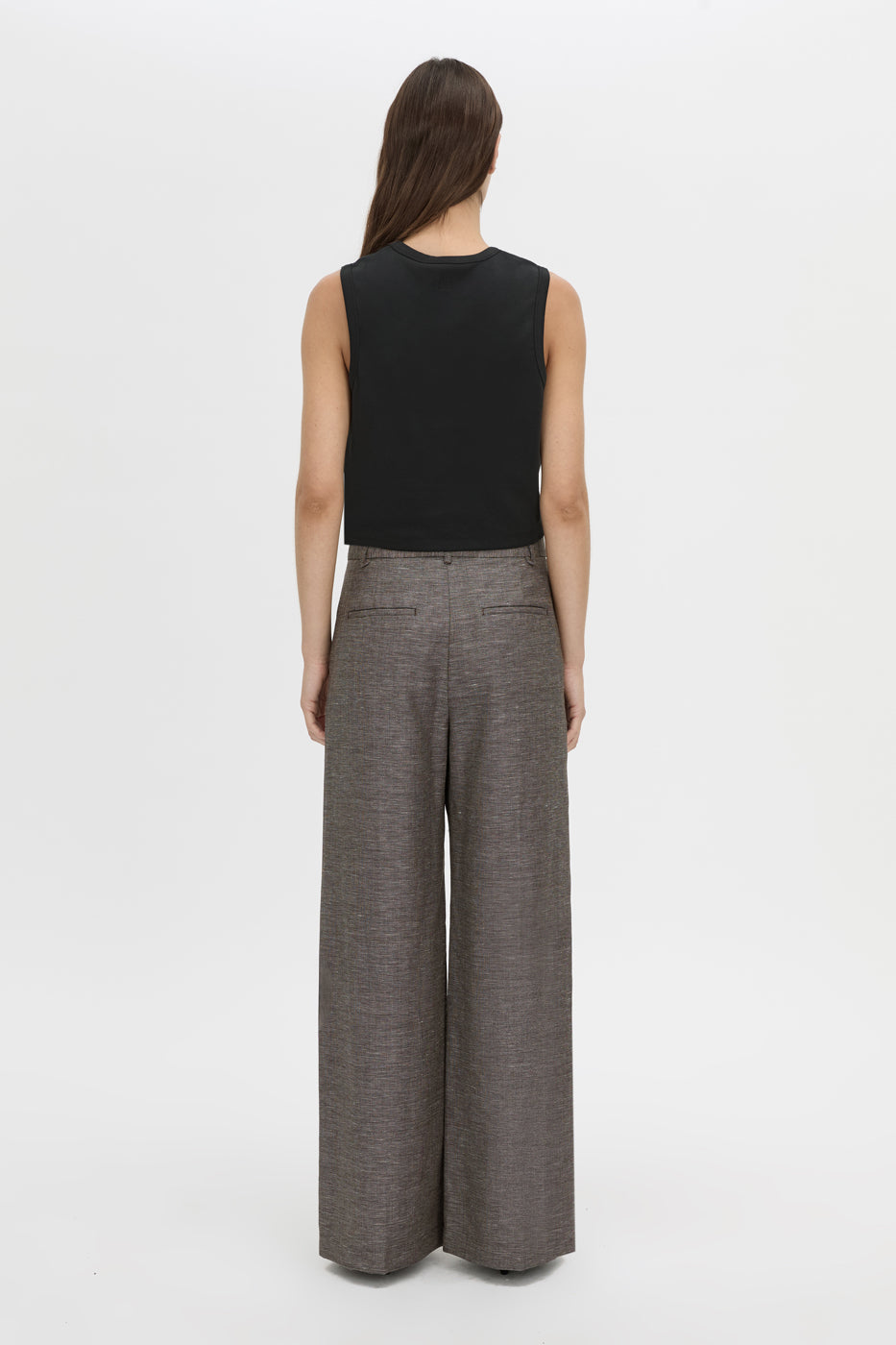 A woman shown from the back wearing a black sleeveless top and high-waisted, wide-leg gray trousers against a plain white studio background.