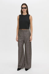 A model stands against a plain light gray studio background wearing a black sleeveless cropped top, wide-leg charcoal gray trousers, and black pointed shoes.