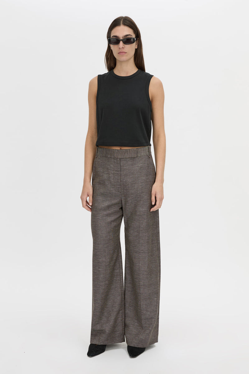 A model stands against a plain light gray studio background wearing a black sleeveless cropped top, wide-leg charcoal gray trousers, and black pointed shoes.