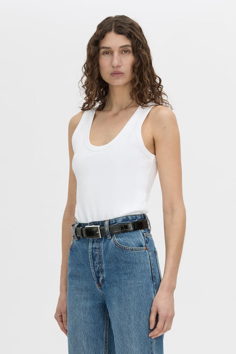A woman modeling a white ribbed scoop-neck tank top paired with blue jeans and a black belt, photographed against a clean white studio background.