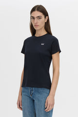 A woman modeling a dark navy blue short-sleeve crewneck t-shirt with a small white embroidered logo on the left chest, paired with blue jeans, against a plain light gray background.