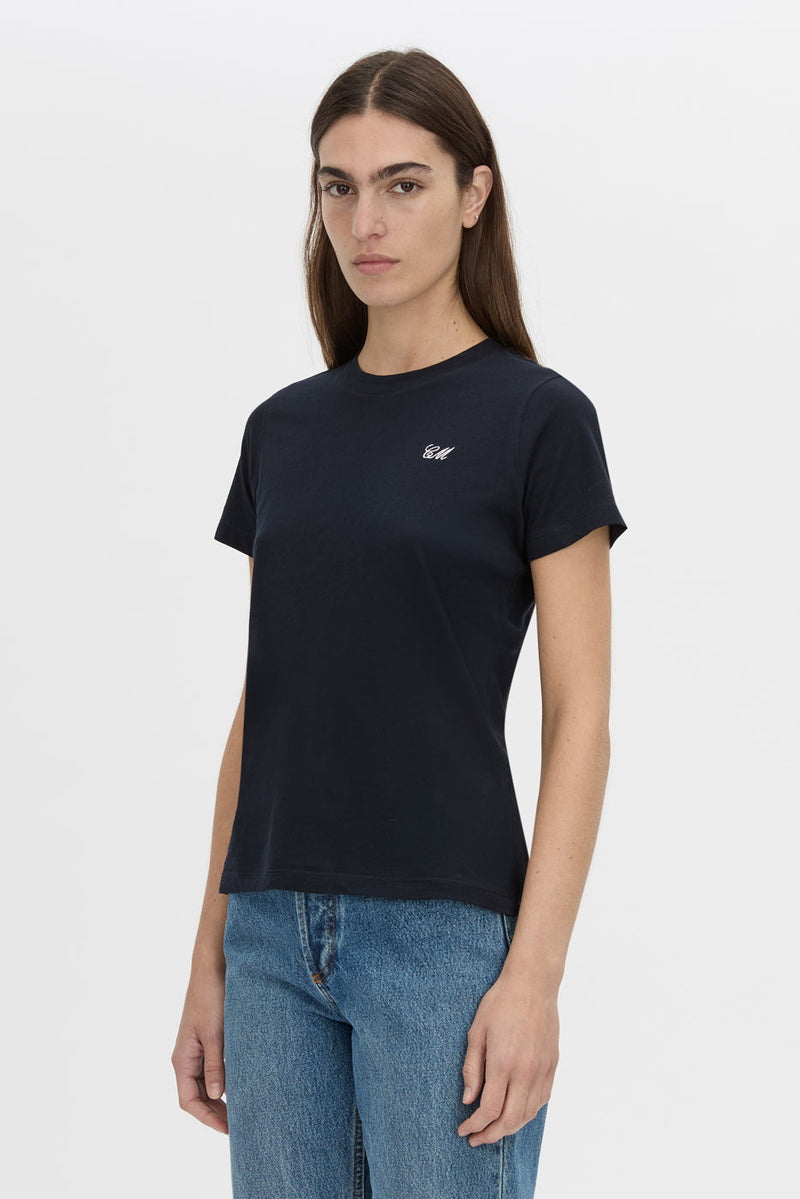 A woman modeling a dark navy blue short-sleeve crewneck t-shirt with a small white embroidered logo on the left chest, paired with blue jeans, against a plain light gray background.