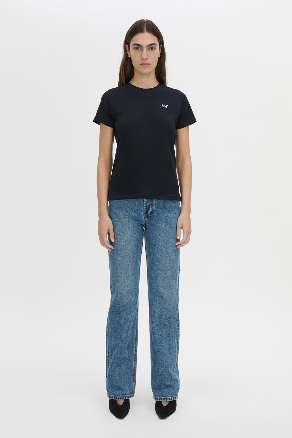 A woman stands against a light gray studio background wearing a black short-sleeve crewneck t-shirt with a small white embroidered logo on the left chest, paired with blue jeans and black pointed shoes.