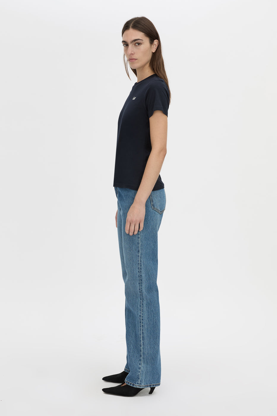 A woman stands in profile wearing a dark navy short-sleeve crewneck t-shirt, light blue straight-leg jeans, and black pointed-toe flats against a plain light gray studio background.