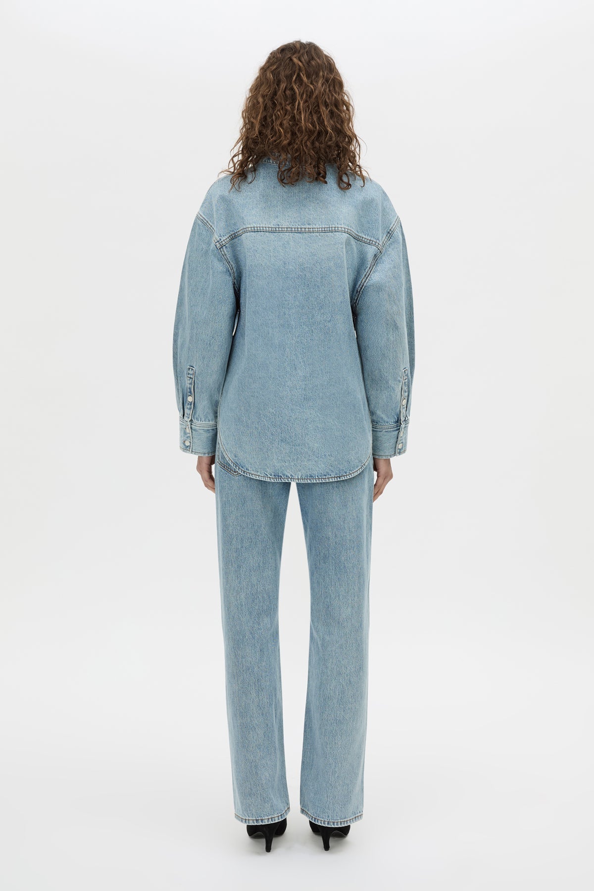 A model viewed from the back wearing a light blue denim jacket and matching wide-leg jeans, paired with black heels, against a plain white studio background.