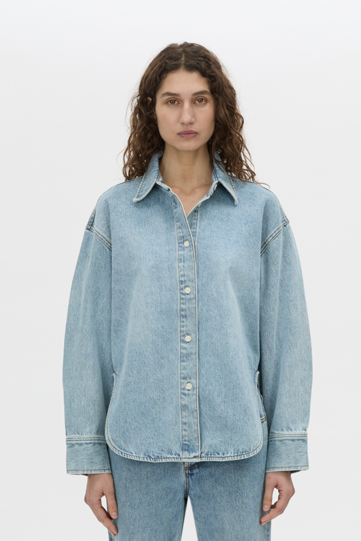 A woman wearing an oversized light blue denim button-up shirt and matching light blue jeans, standing against a plain light gray studio background.