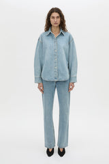 A model stands against a plain white studio background wearing a light blue denim button-up shirt and matching light blue denim jeans with black pointed shoes.
