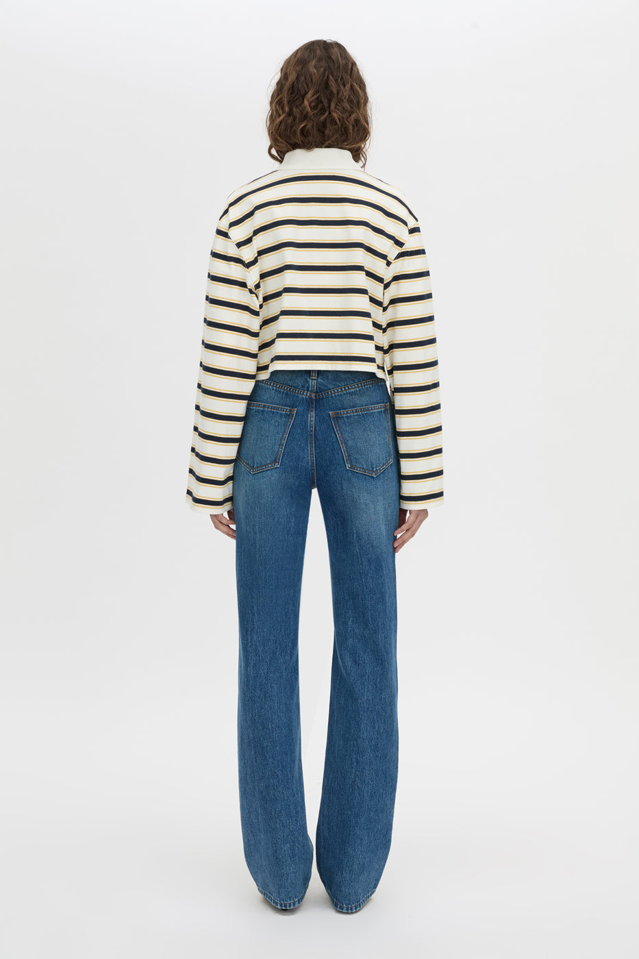 A model viewed from behind wearing a cream and black horizontally striped long-sleeve knit top and blue wide-leg jeans, set against a plain white studio background.