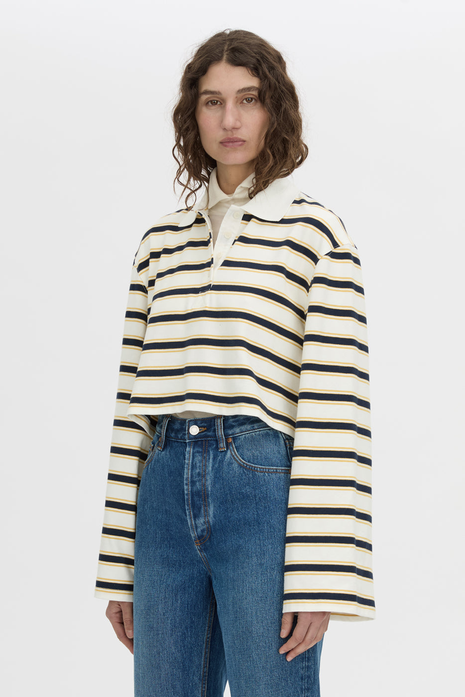 A model wearing a cropped, long-sleeve polo shirt with horizontal navy and cream stripes, tucked into blue jeans, against a plain light gray studio background.