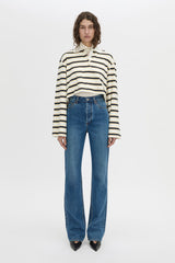 A model stands against a plain white studio background wearing a cream and black horizontally striped long-sleeve cropped polo shirt, blue wide-leg jeans, and black pointed shoes.