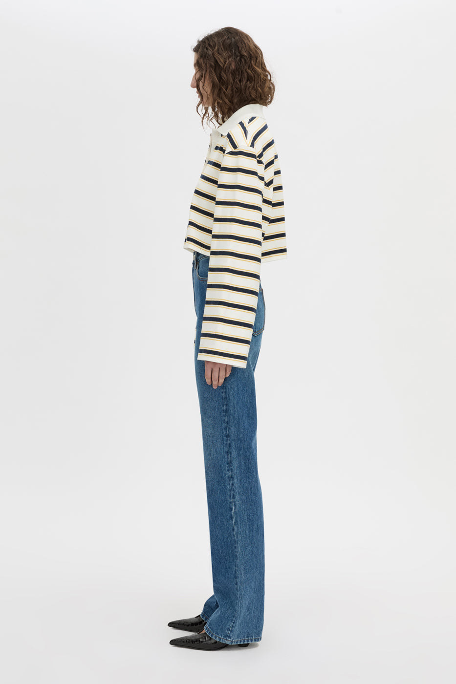 A woman shown from the side profile wearing a cream and black horizontally striped long-sleeve knit top, blue wide-leg jeans, and black shoes, standing against a plain white studio background.