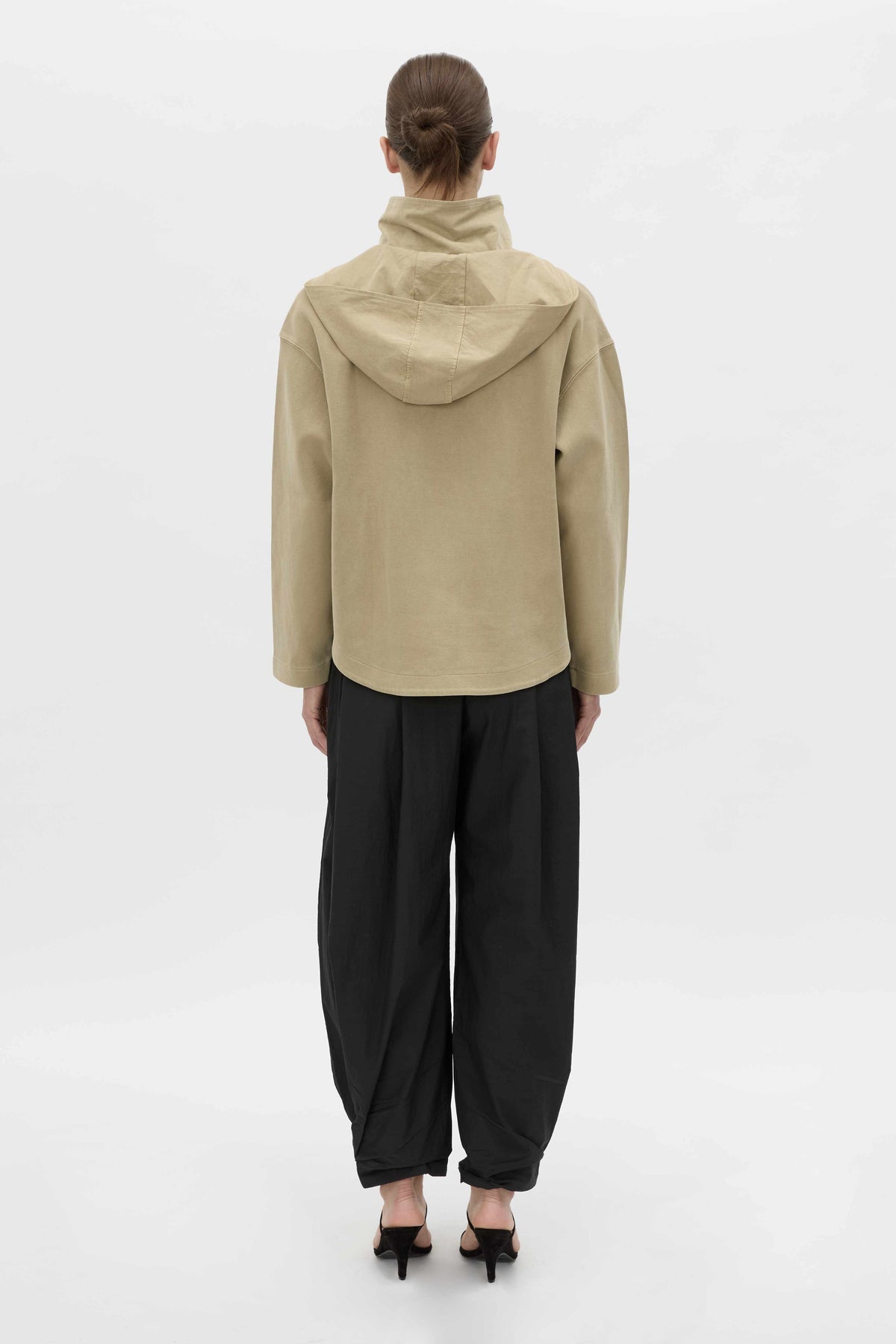 A model viewed from the back wearing a beige hooded jacket and black baggy pants against a plain white studio background.