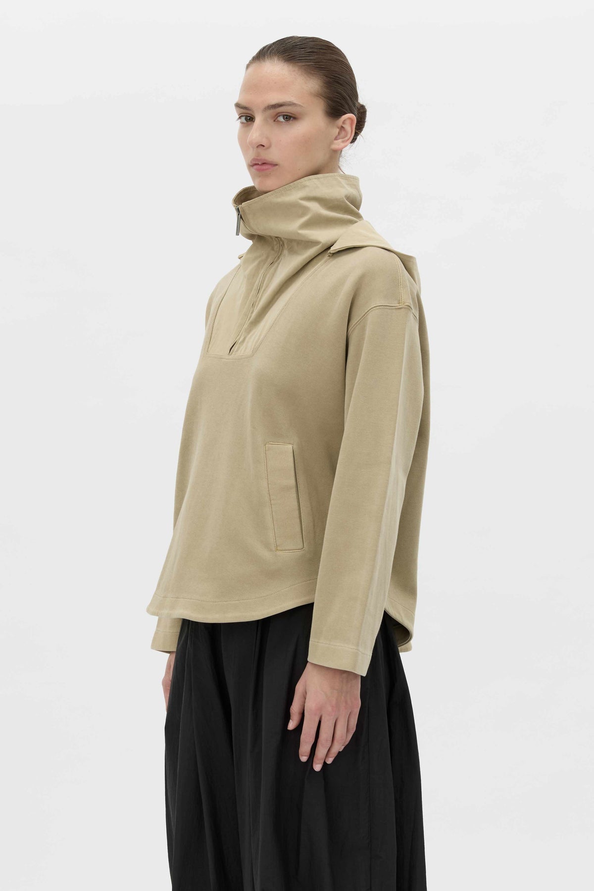 A model wearing a beige high-neck pullover jacket with a side pocket, paired with black wide-leg trousers, photographed against a clean white studio background.