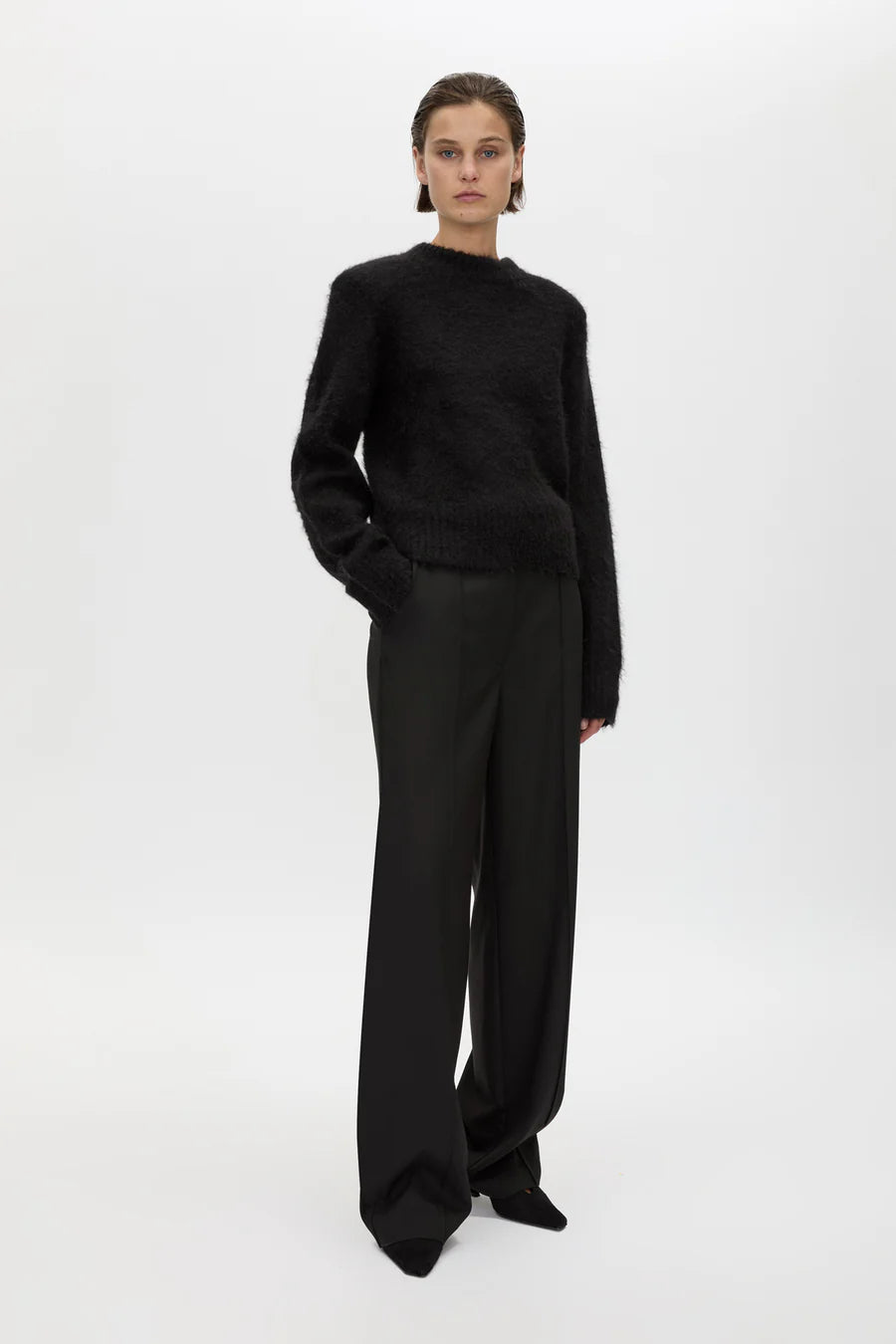 A model stands against a plain light gray studio background wearing a black fuzzy long-sleeve sweater and black wide-leg trousers.