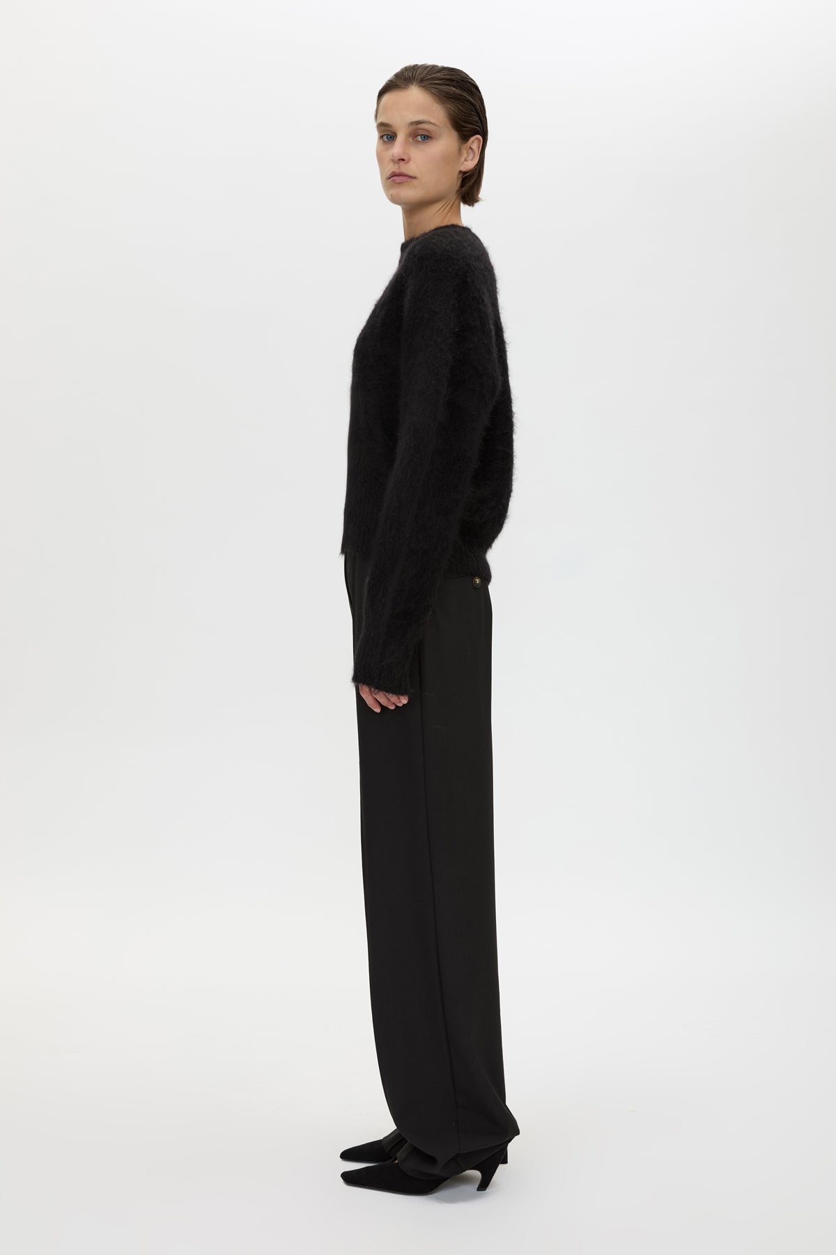 A model is shown in profile wearing a black fuzzy knit sweater and matching black wide-leg trousers against a plain white studio background.