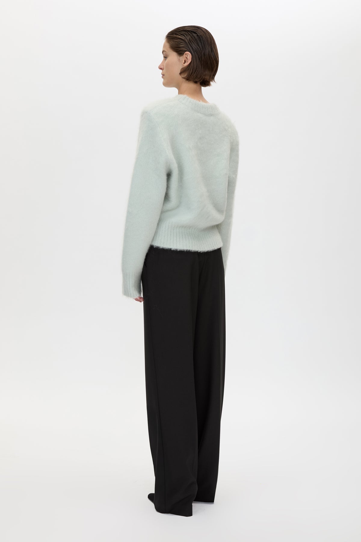 A woman is shown from the back wearing a fuzzy pale sage green sweater and black wide-leg trousers, standing against a plain white studio background.