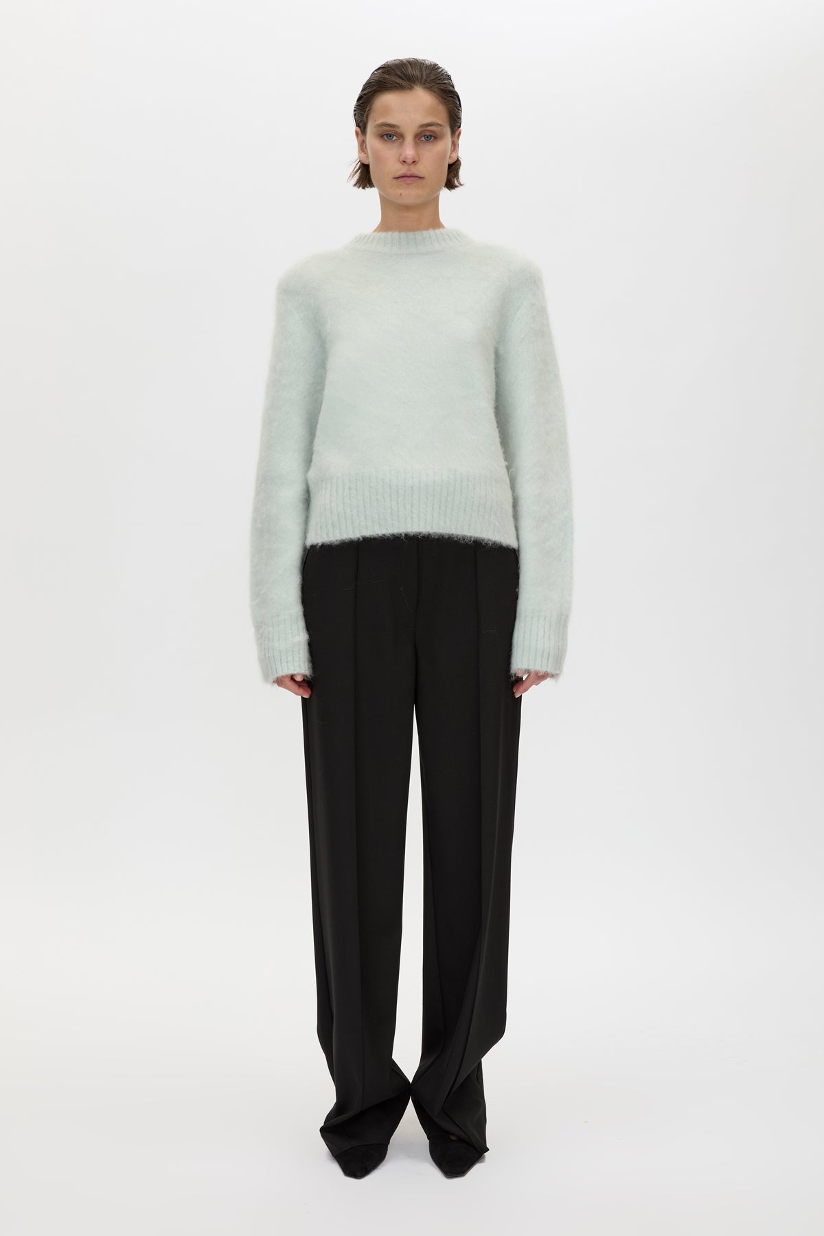 A model stands against a plain white studio background wearing a pale mint green fuzzy knit mock-neck sweater and black wide-leg trousers.