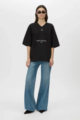 A model wearing an oversized black V-neck t-shirt with white printed text on the front, paired with wide-leg blue jeans and black pointed shoes, standing against a light gray studio background.