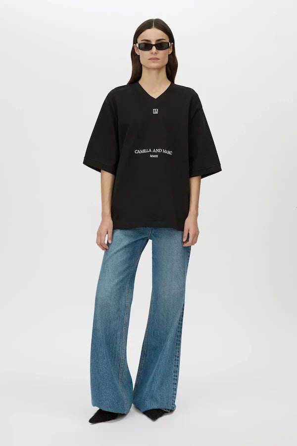 A model wearing an oversized black V-neck t-shirt with white printed text on the front, paired with wide-leg blue jeans and black pointed shoes, standing against a light gray studio background.