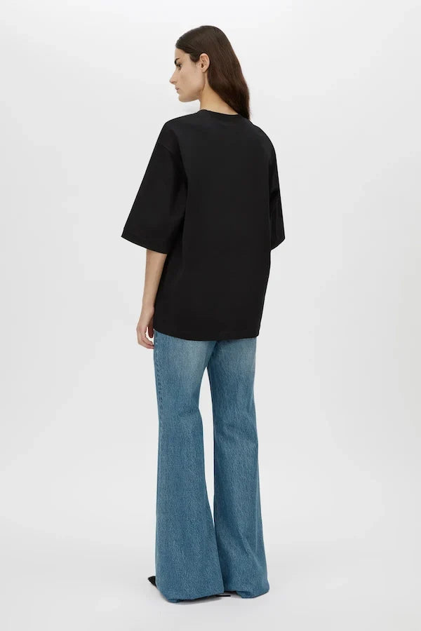 A woman shown from the back wearing an oversized black short-sleeve top and blue flared jeans against a plain light gray studio background.