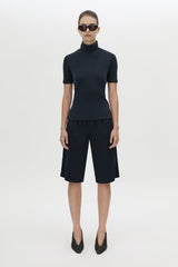 A model stands against a light gray studio background wearing a dark navy short-sleeve mock-neck top and matching dark navy knee-length shorts, with black pointed shoes and sunglasses.